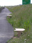 The three benches shown at the side of the cycle track, with a large grass banking behind them, a portrait shot