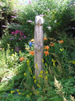 Two tall thin wooden statues closely coupled, surrounded by colorful flowers and plants
